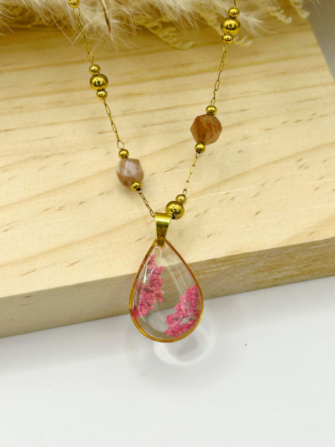 Naszyjnik handmade – przezroczysta, kroplowa zawieszka z żywicy z zatopionymi różowymi suszonymi kwiatami na pozłacanym łańcuszku zdobionym fasetowanymi kamieniami słonecznymi, na miękkim, beżowym tle. Naszyjnik handmade – przezroczysta, kroplowa zawieszka z żywicy z zatopionymi różowymi suszonymi kwiatami na pozłacanym łańcuszku zdobionym fasetowanymi kamieniami słonecznymi, na miękkim, beżowym tle.