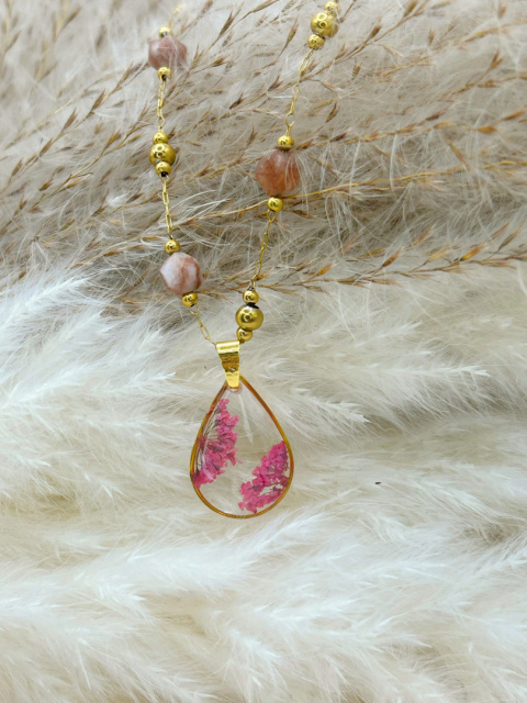 Naszyjnik handmade – przezroczysta, kroplowa zawieszka z żywicy z zatopionymi różowymi suszonymi kwiatami na pozłacanym łańcuszku zdobionym fasetowanymi kamieniami słonecznymi, na miękkim, beżowym tle. Naszyjnik handmade – przezroczysta, kroplowa zawieszka z żywicy z zatopionymi różowymi suszonymi kwiatami na pozłacanym łańcuszku zdobionym fasetowanymi kamieniami słonecznymi, na miękkim, beżowym tle.