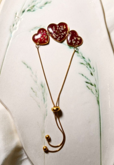 Ręcznie robiona bransoletka z żywicy w kształcie serc z białymi kwiatami, w oprawie złotej stali,  na dekoracyjnej powierzchni.